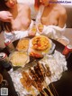 Two naked women sitting at a table with food and drinks.