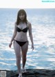 A woman in a black bikini standing on a rock by the ocean.